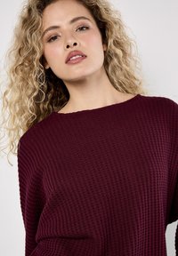 Vrouw met krullend blond haar, gekleed in een donkerpaarse gebreide trui, die iets omhoog kijkt met een neutrale uitdrukking.
