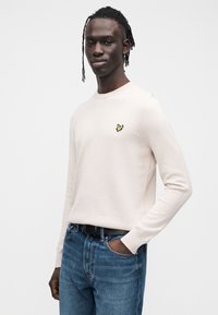 Ljusbeige tröja i mjukt tyg med långa ärmar och rund halsringning. Har en liten gul fågel-logga på vänstra bröstet. Bärs med blå jeans.