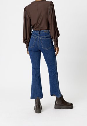 Blusa larga de manga larga marrón con hombros abullonados, combinada con vaqueros de mezclilla azul acampanados. La modelo lleva botas de tobillo marrón oscuro. Diseño minimalista.