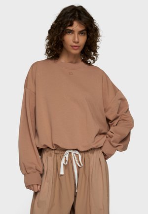Junge Frau mit lockigem Haar trägt einen lockeren braunen Sweatshirt und passende Hose mit weißem Kordelzug vor einem einfarbigen Hintergrund.
