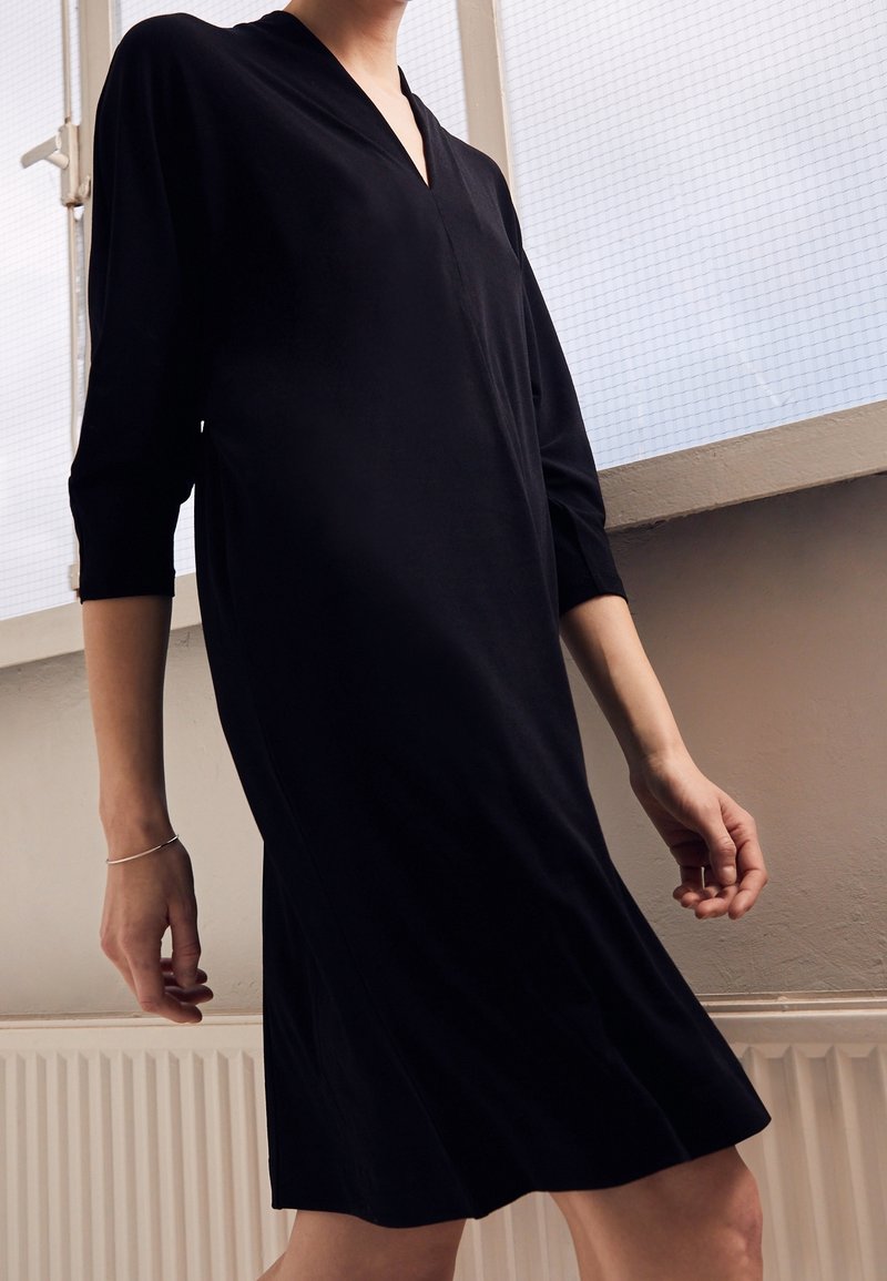 Personne portant une robe noire à manches longues debout à l'intérieur près d'une fenêtre avec un bracelet au poignet gauche.