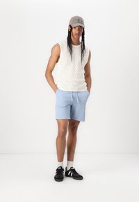 Top sans manches crème en tricot, shorts bleu clair, baskets noires avec accents blancs, casquette grise, se tenant sur un fond blanc uni.