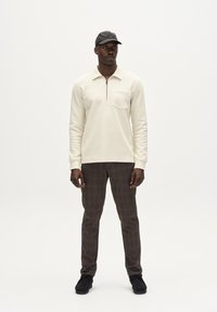 Pullover beige con mezza zip e tasca, abbinato a pantaloni a quadri marroni e scarpe nere. Modello con cappellino nero. Sfondo neutro.