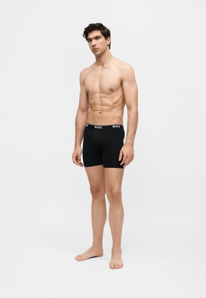 Mladi muškarac s fit fizičkom građom stoji bos, nosi crne bokserice marke BOSS na jednostavnoj svijetloj pozadini.