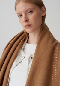 Sciarpa di lana marrone appoggiata su una maglietta bianca e una giacca leggera, caratterizzata da una texture liscia e un motivo sottile lungo il bordo.