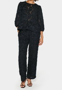 Kobieta nosząca czarną, przezroczystą bluzkę peplum z długim rękawem oraz pasujące, teksturowane szerokie spodnie z czarnymi, odkrytymi czółenkami na wysokim obcasie.