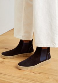 Schwarze Wildleder-Stiefeletten mit einem strukturierten blauen elastischen Einsatz und einer gerippten beigen Sohle. Getragen mit weißen weit geschnittenen Hosen auf einem Holzboden.