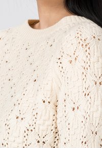 Pull en laine couleur crème avec un design texturé comportant des motifs ajourés. Les manches ont un col rond et une bordure en côtes.