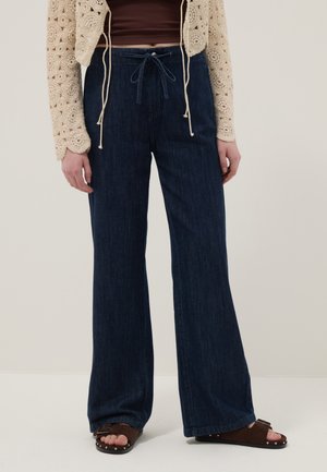 Femme portant un pantalon en jean bleu foncé à larges jambes avec une taille à cordon, des sandales marron à clous, et un cardigan en crochet beige.