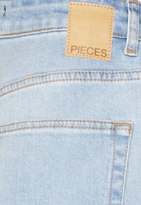 Jeans de ganga azul claro com uma etiqueta de couro visível rotulada "PIECES." Apresenta costuras e um design de bolso traseiro. Textura suave, estilo casual.