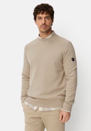 Beige strikket sweater med rund halsudskæring og ribbede manchetter. Bæres over en ternet skjorte. Har et lille sort logo på venstre ærme.