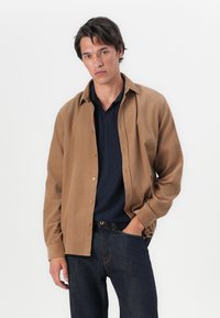 Chemise brune à boutons avec un col classique, portée sur un polo en laine bleu marine. Présente des manches longues et une coupe décontractée. Jeans en denim foncé.