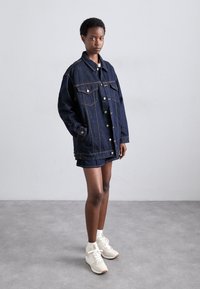 Giacca in denim di colore blu scuro, dal fit oversize, con due tasche sul petto, bottoni a pressione e cuciture gialle. Abbinata a una mini gonna in denim.