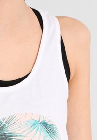 Biały, bezrękawnik z grafiką liścia palmy w turkusie i różu, połączony z czarnym sportowym biustonoszem pod spodem.