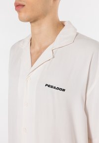 Man draagt een lichtbeige overhemdbloes met een open kraag en "PEGADOR" in zwarte letters geborduurd op de linkerborst.