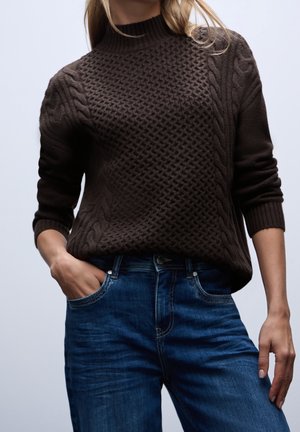 Personne portant un pull en maille texturée marron foncé rentré dans un jean en denim bleu, une main dans la poche, sur un fond uni.