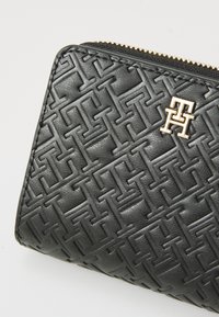 Portefeuille en cuir noir avec un motif géométrique en relief, présentant un accent logo en ton doré et une fermeture éclair le long du bord supérieur.