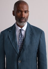 Blaugrüner Leinenblazer mit Kerbkragen, einem braunen Knopf und einer Brusttasche. Kombiniert mit einem hellblauen Hemd und einer gemusterten marineblauen Krawatte.