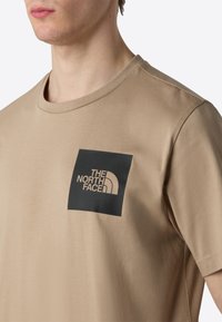 Beige kortærmet t-shirt lavet af blødt stof, der har et sort rektangulært logo-patch på brystet med hvid tekst og buet design.