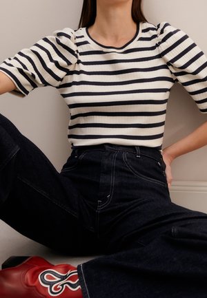 Vrouw draagt een zwart-wit gestreept topje met korte mouwen, donkere hooggetailleerde spijkerbroek en rode laarzen met een decoratief patroon.