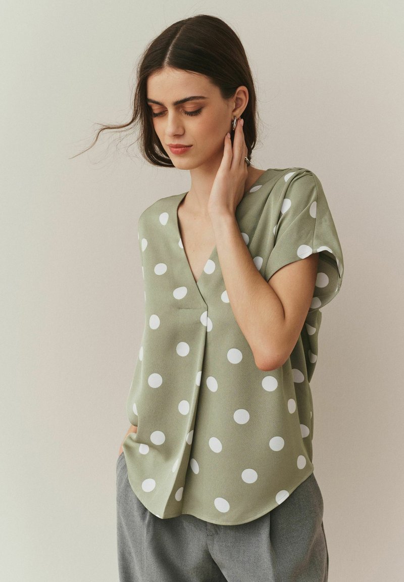 Lichtgroene blouse met grote witte polka dots. Heeft een diepe V-hals en korte mouwen, gemaakt van een gladde stof met een losse pasvorm.