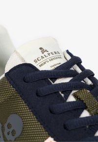 Heren lifestyle sneaker met marineblauwe en olijfgroene stof, textuuraccenten, een witte tonglabel en een doodskoplogo.