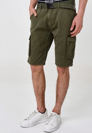 Homme portant un short cargo vert olive avec poches avant et latérales, une ceinture verte, une ceinture de sous-vêtements noire et des baskets blanches sur un fond uni.