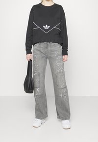 Person wearing black Adidas sweatshirt, gray paint-splattered cargo jeans, white sneakers, holding black handbag.