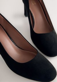 Coppia di scarpe da donna con tacco alto in suede nero con plantari marroni, punte arrotondate e tacchi a blocco su sfondo chiaro.