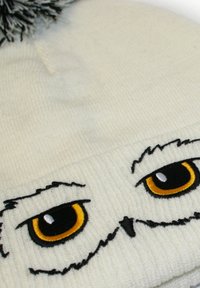 White knitted beanie with a textured finish, featuring embroidered owl eyes in black, yellow, and white, and topped with a black pom-pom.