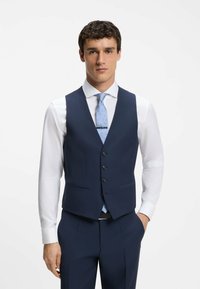 Homme portant un gilet de costume bleu marine sur une chemise blanche avec une cravate bleu clair à motif et une pince à cravate, debout avec une main dans la poche.