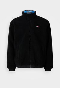 Black fleece jacket with a high collar, front zipper, elastic cuffs, and hem; features a blue and plaid lining inside. Small logo detail on chest.