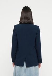 Marineblazer laget av glatt stoff, med skreddersydd design, ventilerte bak, og knappede mansjetter, båret over et denimskjørt.