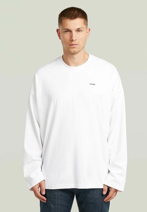 Man met kort haar die een effen wit shirt met lange mouwen draagt met kleine "G-STAR" tekst op de borst en donkere jeans tegen een effen achtergrond.