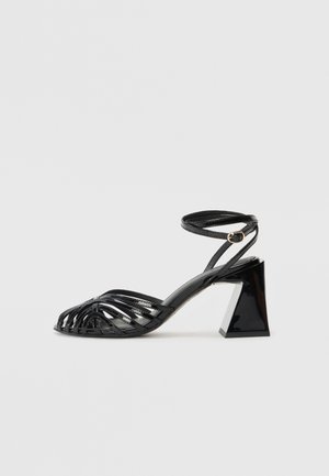 Sandales à talons noires à lanières avec un design tissé, un talon carré épais, et une bride de cheville avec une boucle dorée.