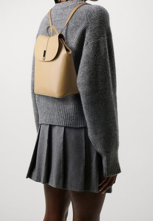 Ryggsäck i lyst tanläder med rundad klaff, justerbara remmar och elegant metallbeslag, kombinerad med en grå stickad tröja och plisserad kjol.