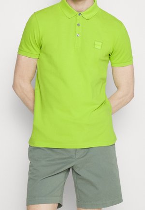 Mand iført en lysegrøn kortærmet poloshirt med knapper og grå shorts, stående med hænderne på ryggen.