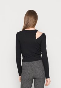 Schwarzes Langarmoberteil mit taillierter Passform, versehen mit einem Ausschnitt an einer Schulter, aus dehnbarem Stoff. Kombiniert mit gemusterten grauen Leggings.