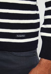 Granatowy i biały sweter z paskami wykonany z dzianiny, z małą czarną metką z napisem "Armor Lux" na dolnym brzegu.