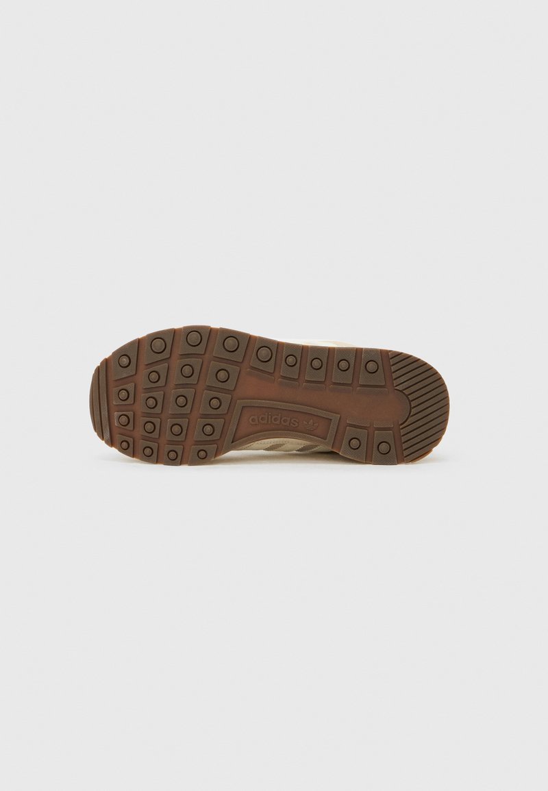 Semelle extérieure d'une chaussure Adidas beige avec une semelle en caoutchouc marron texturée, dotée de crampons circulaires et du logo Adidas au centre.