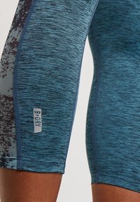 Närbild av blå strukturerade sportshorts med ett mönstrat sidoflik och en "B-Dry"-etikett nära fållen på en persons lår.