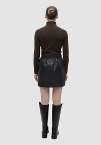 Maglione aderente marrone a collo alto, mini gonna nera in pelle con vita elastica e stivali neri alti fino al ginocchio, visti da dietro su uno sfondo grigio.