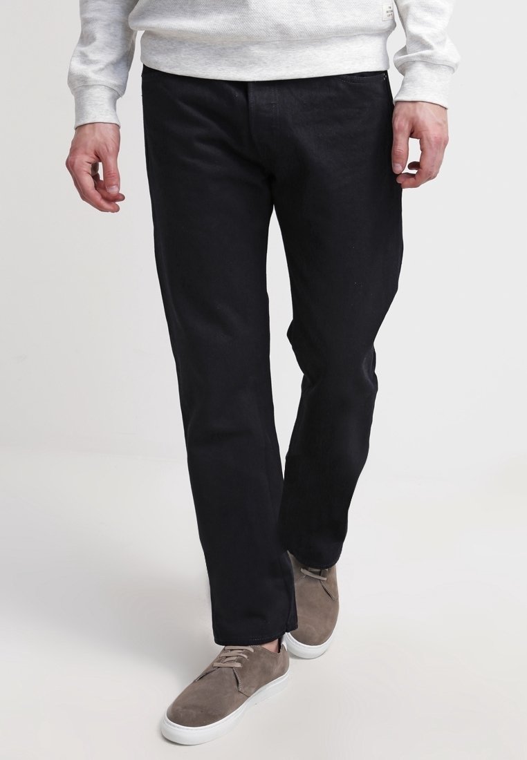 Homme portant un pantalon noir droit, des baskets en daim beige avec des semelles blanches et une chemise à manches longues gris clair, marchant vers l'avant.