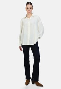 Hvid skjorte med knapper, krave og lange ærmer, stylet med mørke flared jeans og brune hæle støvler. Glat stoftekstur.