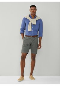 Herreoutfit bestående af en blå knappet skjorte, grå shorts, brunt vævet bælte og beige slip-ons. En sweater er draperet afslappet over skuldrene.