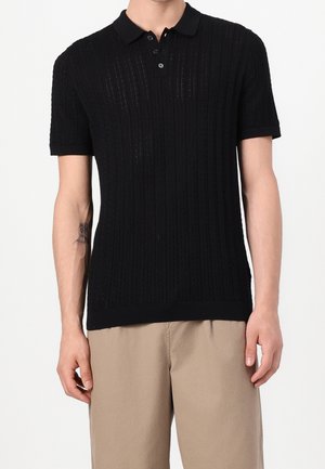 Mann trägt ein schwarzes, kurzärmeliges Strick-Poloshirt mit Knöpfen, kombiniert mit beigen Hosen vor weißem Hintergrund.