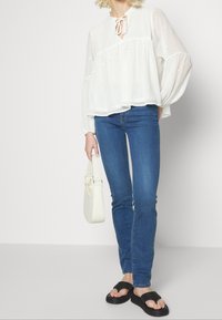 Žena v bílé průhledné halence s dlouhým rukávem, modrých úzkých džínách, černých sandálech a držící malé bílé kabelky.