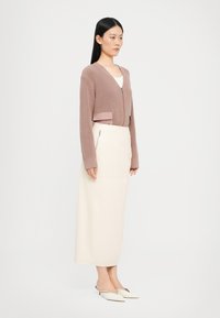 Lys rosa strikket cardigan med v-utringning og en satenglomme, kombinert med en kremfarget midiskjørt med glidelås på siden. Hvite slip-on sko.