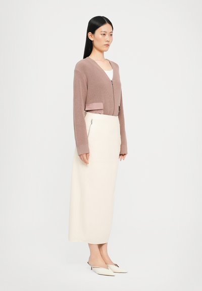 Lys rosa strikket cardigan med v-utringning og en satenglomme, kombinert med en kremfarget midiskjørt med glidelås på siden. Hvite slip-on sko.