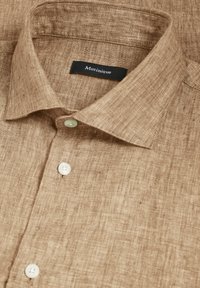 Camicia di lino marrone chiaro con colletto classico e cinque bottoni. Presenta un tessuto testurizzato e un'etichetta nera con la scritta "Matinique."